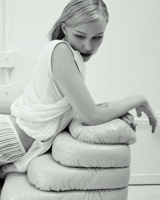 Monochrome image of a blonde woman in a white top and striped bottoms reclining on a stacked cushion, with pale skin and dark lipstick.