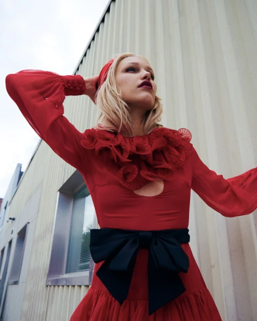 Fashionable blonde woman in a red dress and headscarf strikes a pose against a metal building in an outdoor setting.