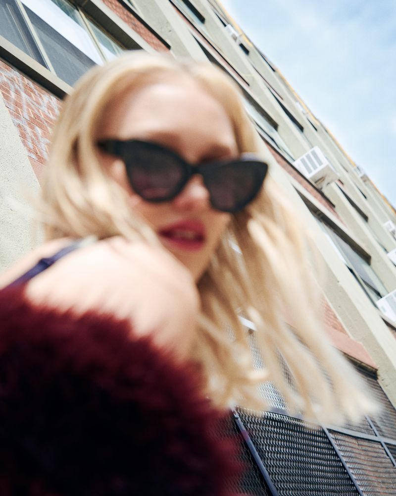Woman with blonde hair wears sunglasses outdoors near a red brick building on a sunny day in an upward angle shot.