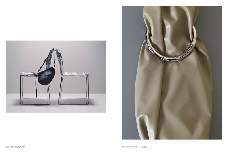 Studio product shot: a black leather bag hanging on a wireframe chair and a silver necklace on a beige draped fabric.