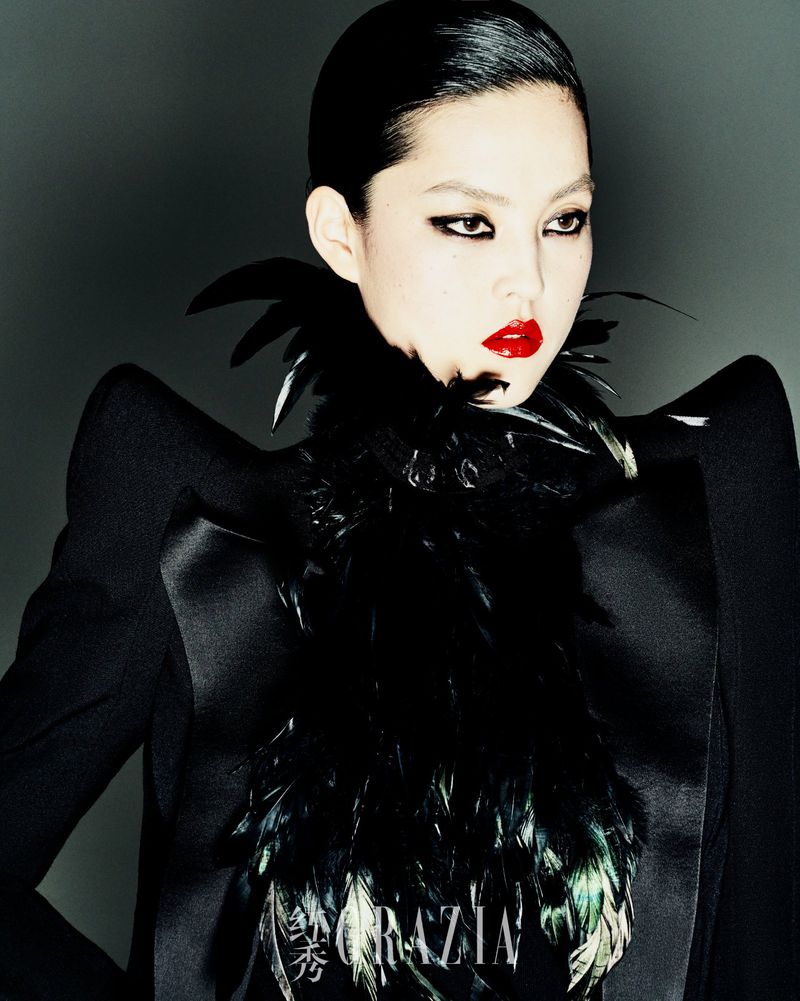 A striking portrait of an Asian woman with red lips, dramatic eye makeup, and feathered collar, wearing a dark jacket against a grey background.