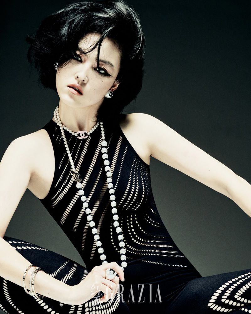 Elegant woman in patterned jumpsuit and layered pearl necklaces poses in a fashion-editorial shot against a dark studio background.
