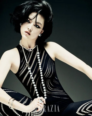Elegant woman in patterned jumpsuit and layered pearl necklaces poses in a fashion-editorial shot against a dark studio background.