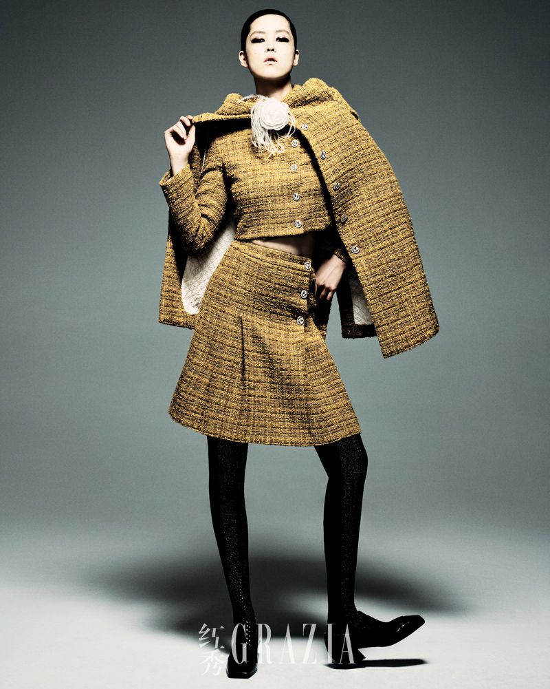 Full-length shot of an Asian fashion model in a yellow tweed two-piece outfit with black tights and shoes, posing in a studio.