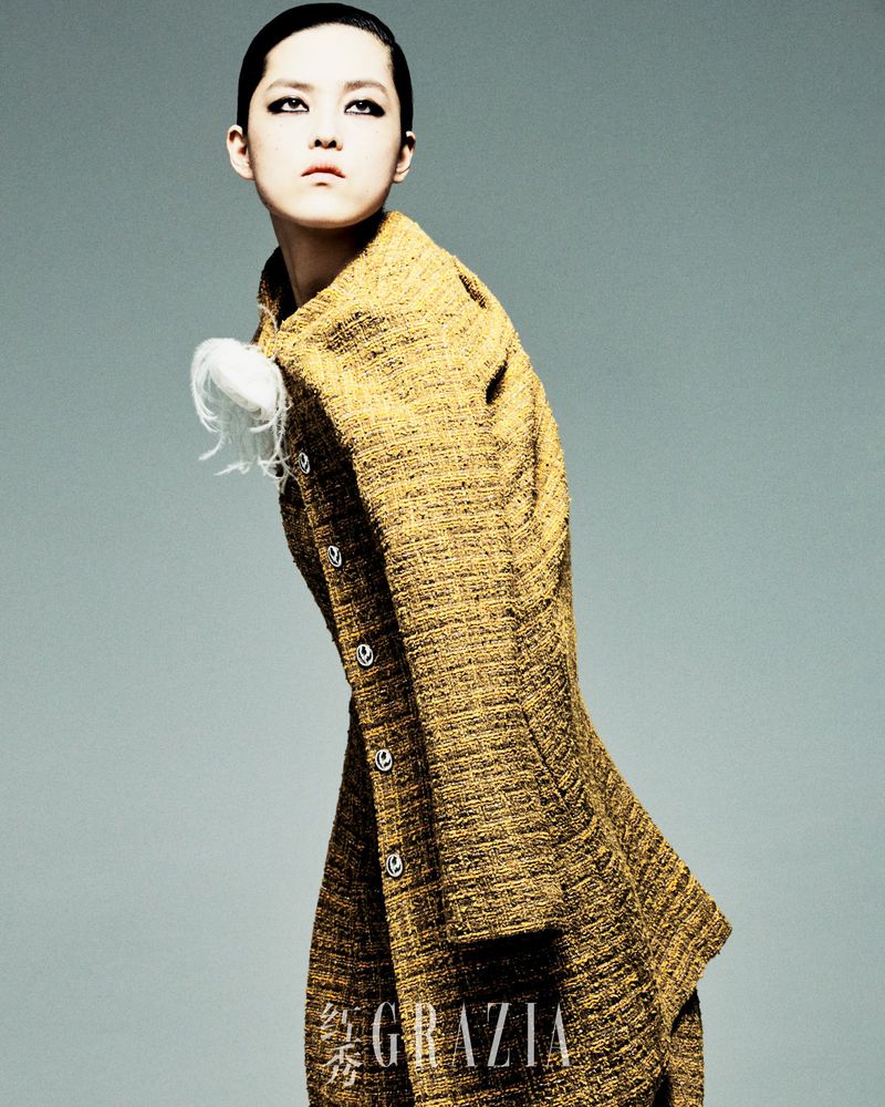 Woman in patterned yellow coat poses in front of grey background. She has short hair and is wearing dramatic dark eye makeup.