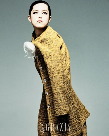 Woman in patterned yellow coat poses in front of grey background. She has short hair and is wearing dramatic dark eye makeup.