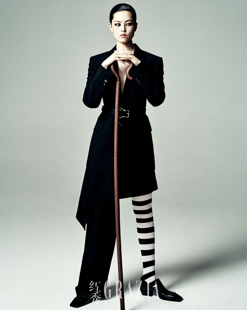 A stylish Asian woman in a black belted jacket holds a cane, standing against a neutral backdrop, showcasing a bold fashion statement.