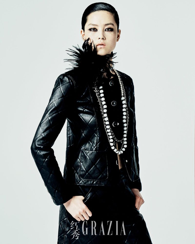 An Asian woman poses in a black quilted jacket and skirt, adorned with a feathered collar and pearl necklace, against a pale backdrop.