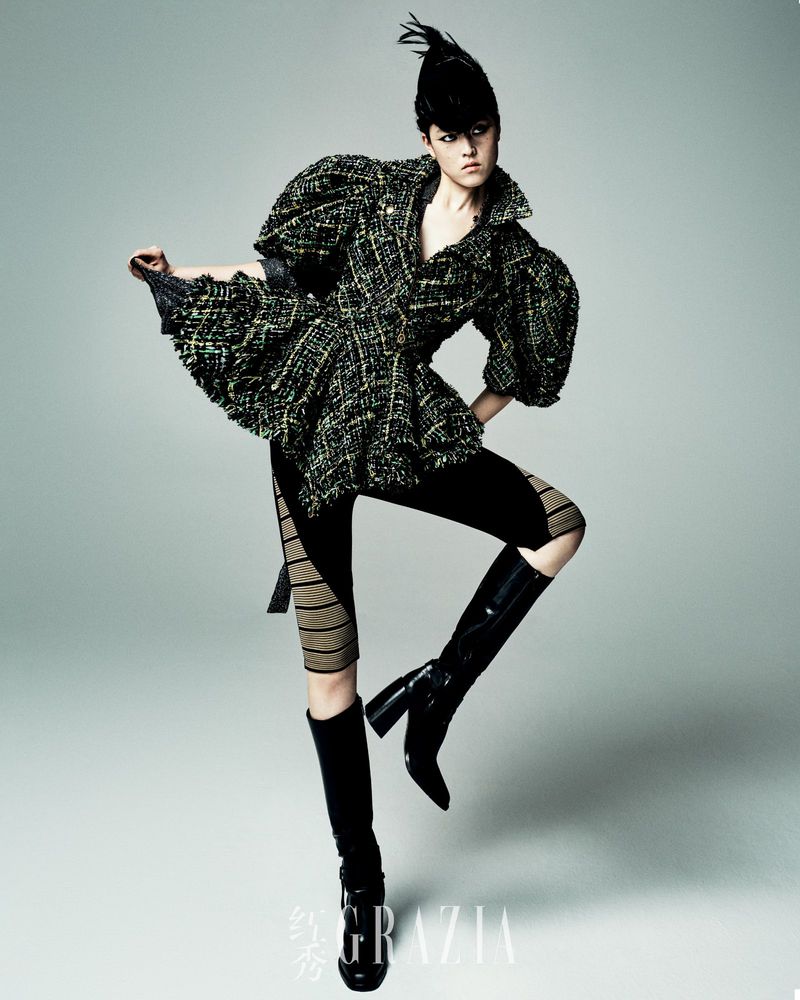 A fashion model strikes a dynamic pose in a textured tweed jacket, leggings, and knee-high boots against a gradient grey backdrop.