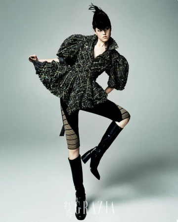 A fashion model strikes a dynamic pose in a textured tweed jacket, leggings, and knee-high boots against a gradient grey backdrop.