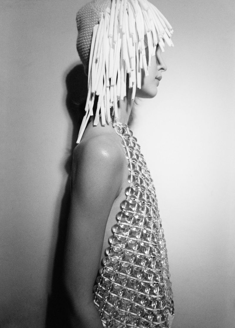 Side profile portrait of a woman in a knit cap with fringe, wearing a shimmering, open weave dress against a neutral wall.