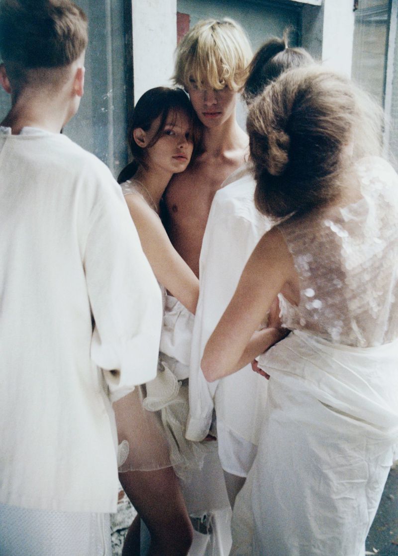 Several young adults in a close group, wearing textured white clothing in a gritty urban setting, lit with diffused natural light.
