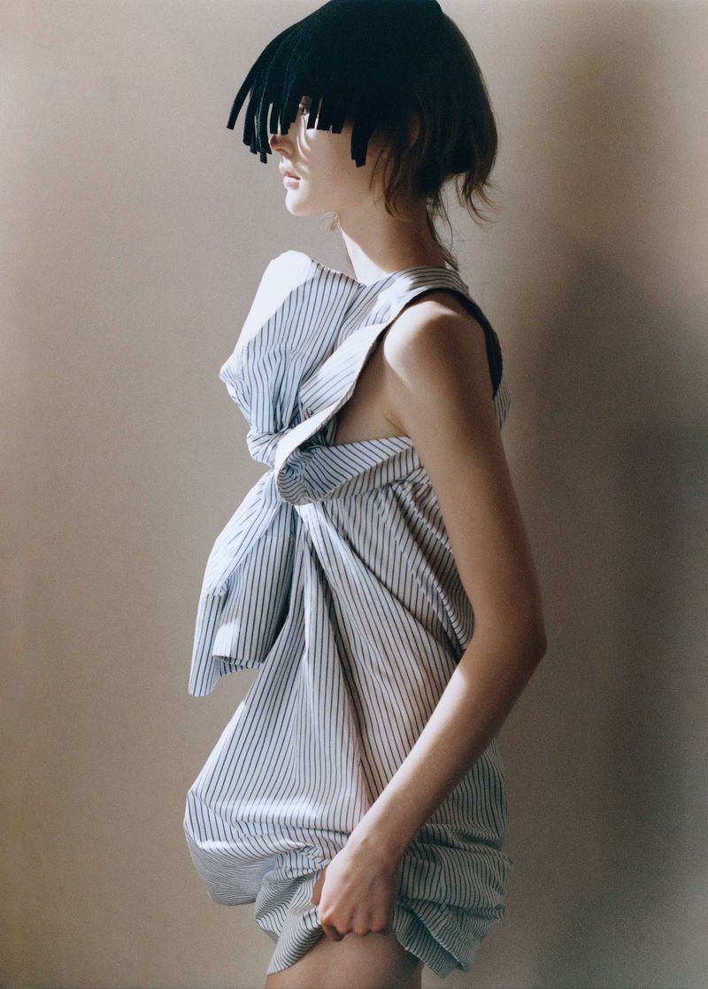 A woman in a striped dress poses in a studio setting. She is wearing a dark wig with short fringe. Soft, diffused lighting.