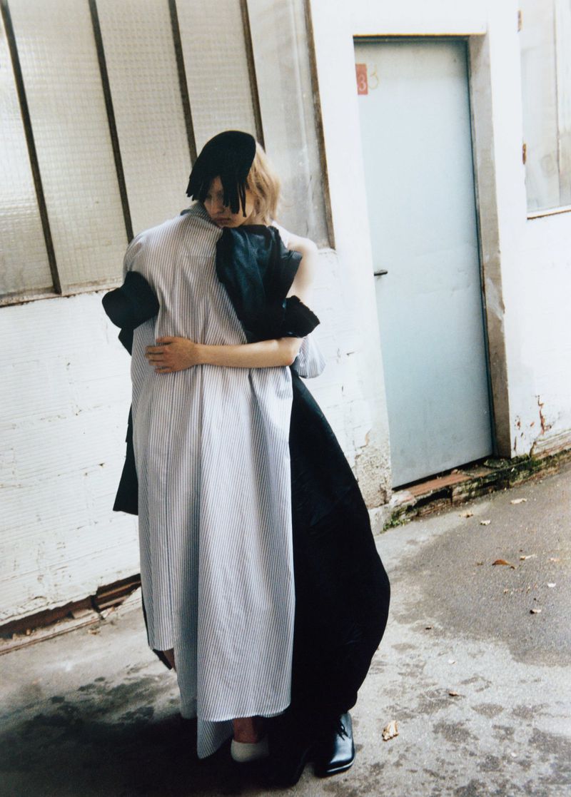 Two people embrace outdoors, one in a long-sleeved striped dress, the other in a black gown, by a concrete wall and metal door.