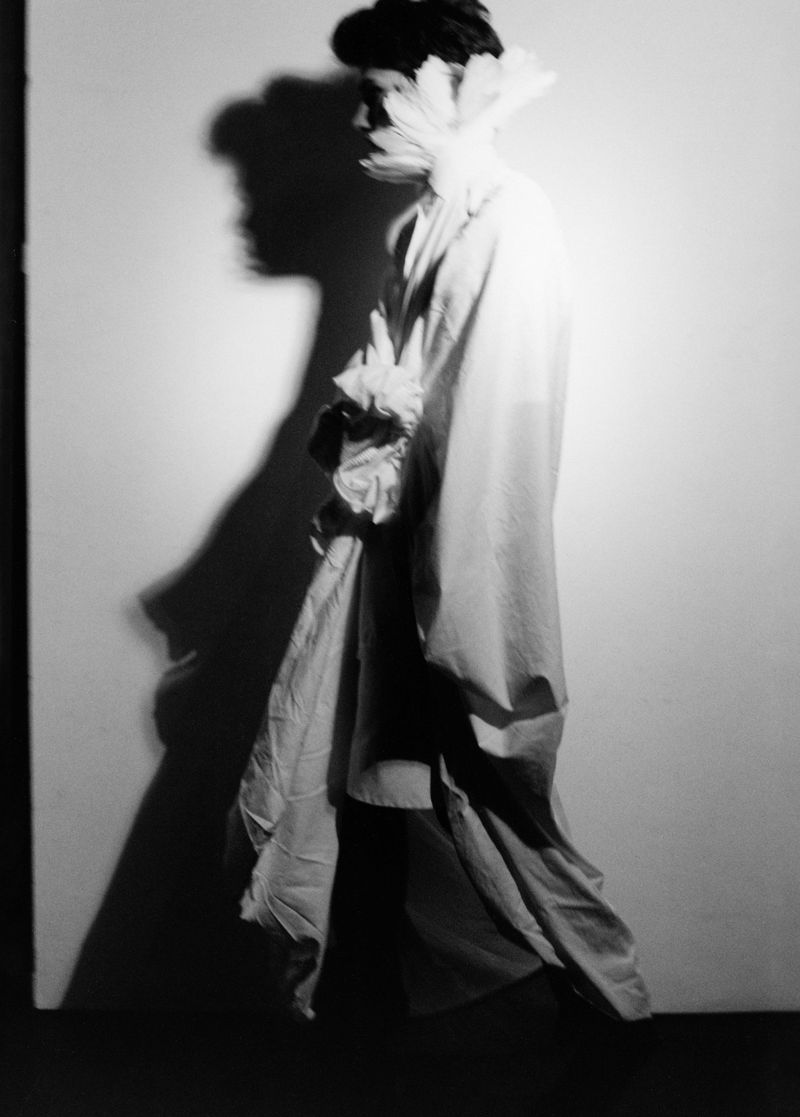 Monochrome portrait of a person in a white gown and feathered neckpiece with a dark, shadowy silhouette against a white background.