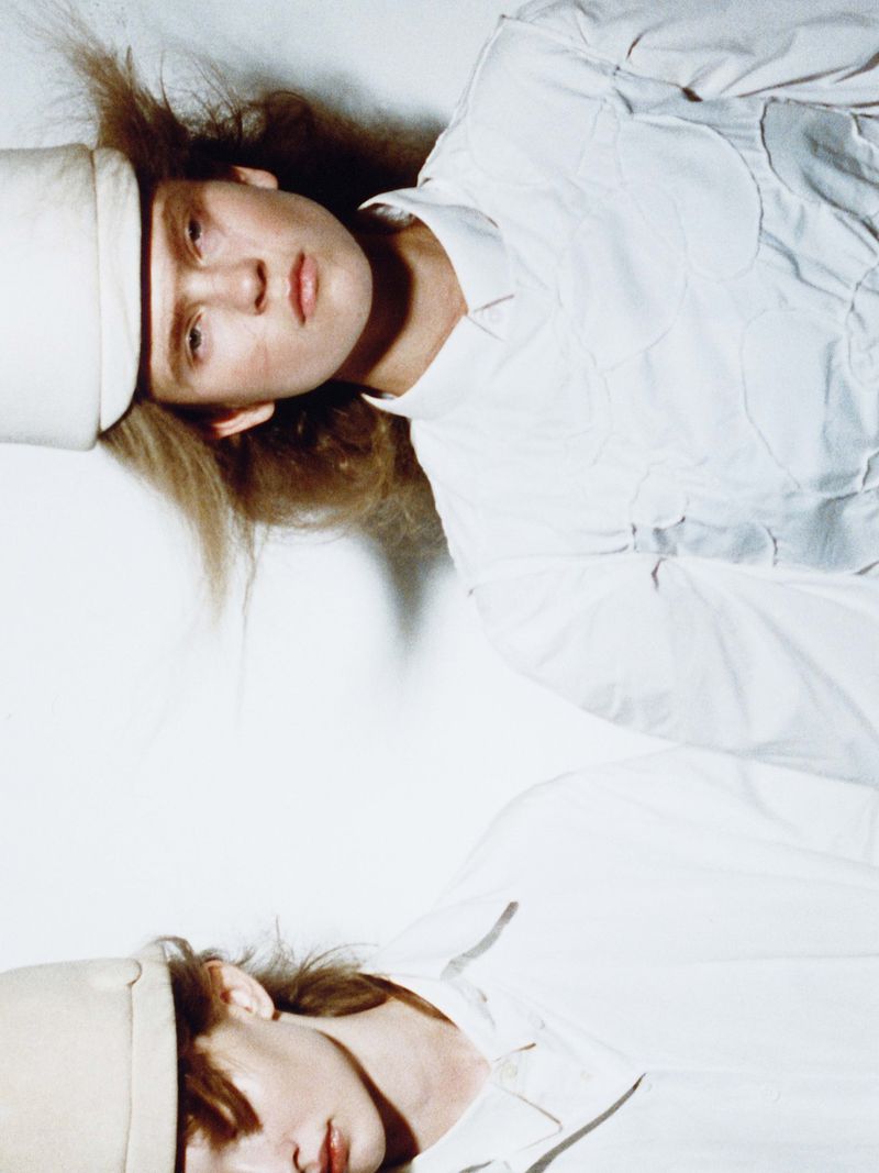 Conceptual fashion shot of two models lying on a white surface, wearing matching white shirts and hats with a bright, even lighting.