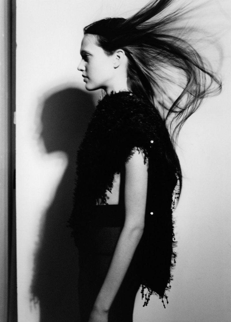 Profile portrait of a model with long dark hair blowing in motion, wearing a black textured sleeveless top against a white wall.