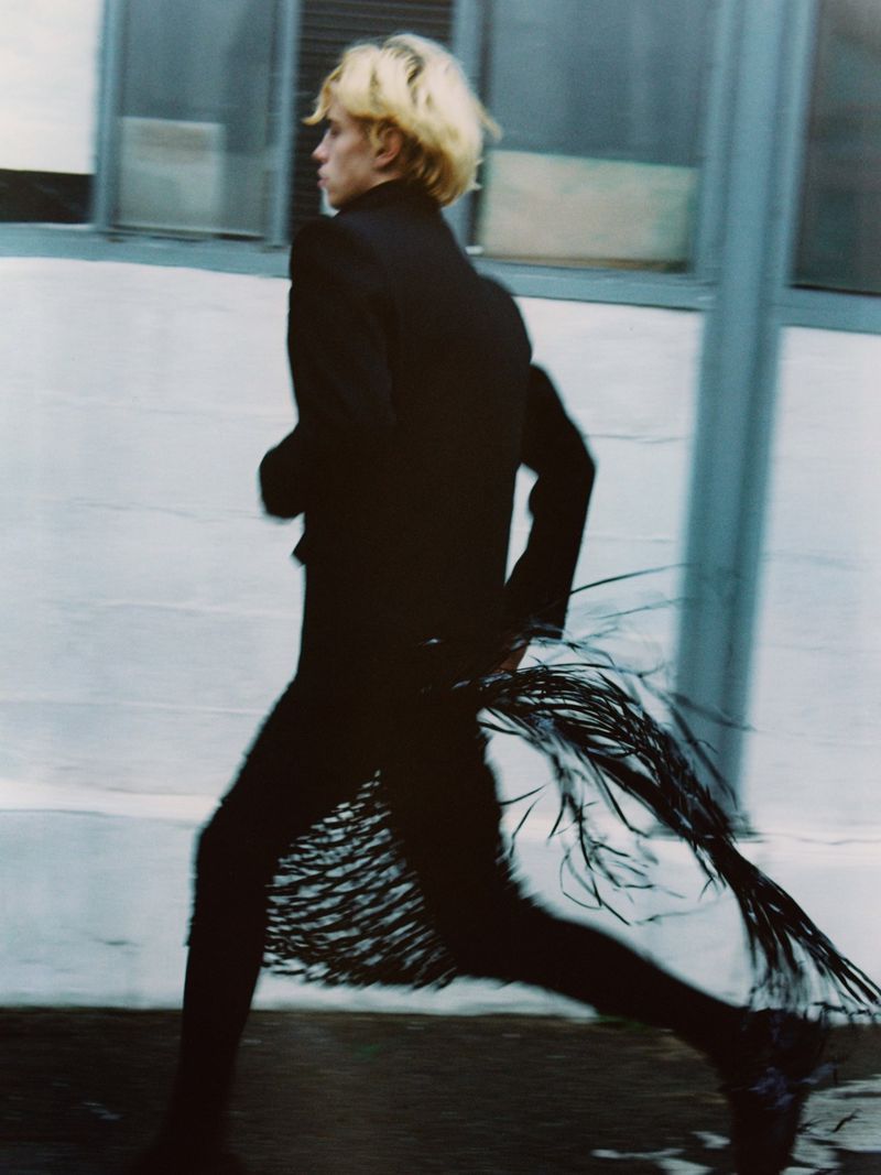 Fashion photo of a blond adult model in black fringed suit running outdoors against a concrete wall on a city street in a fashion editorial.