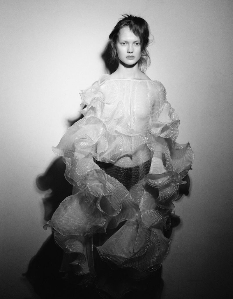Monochromatic portrait of a young woman wearing a ruffled top, posing in front of a textured studio backdrop. Fashion photography.