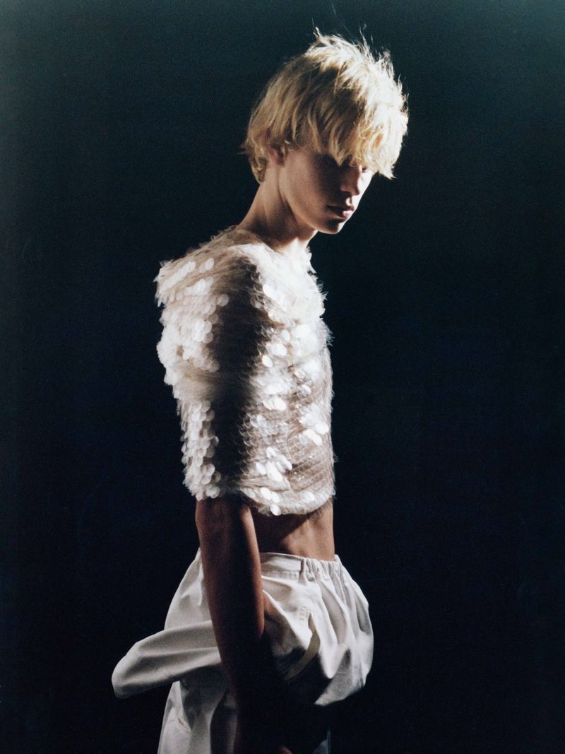 Androgynous model poses in white top and pants against dark backdrop in studio lighting, side view, for fashion editorial photography.