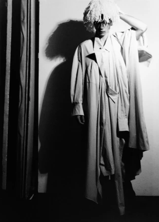 Model wearing a textured white hat and layered coat casting a dark shadow on a wall in a black and white editorial photograph.