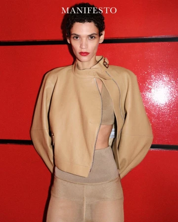 Fashion shot of a slender woman in a beige jacket and pants standing against a red wall with black stripes in a studio setting.