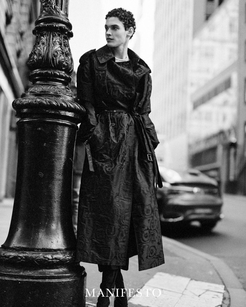 Black and white shot of a man in a patterned trench coat leaning against a decorative street lamp in an urban setting with a car in the background.