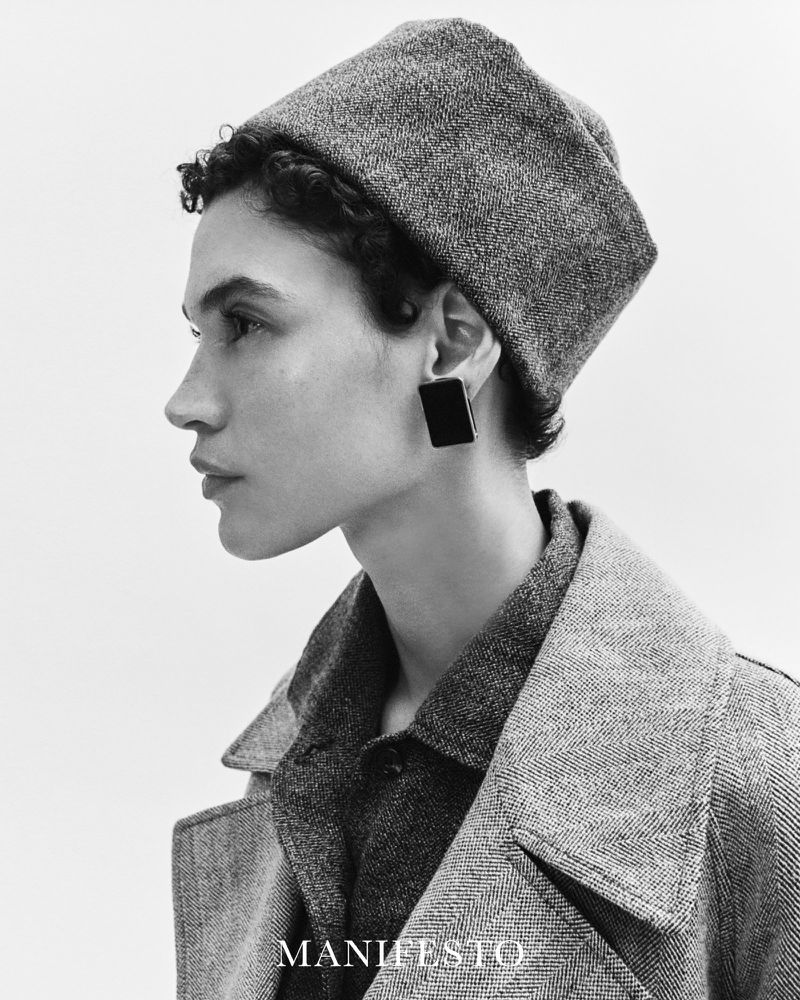 Black and white portrait of a female model wearing a textured hat and coat with a serious expression, emphasizing fashion and style.