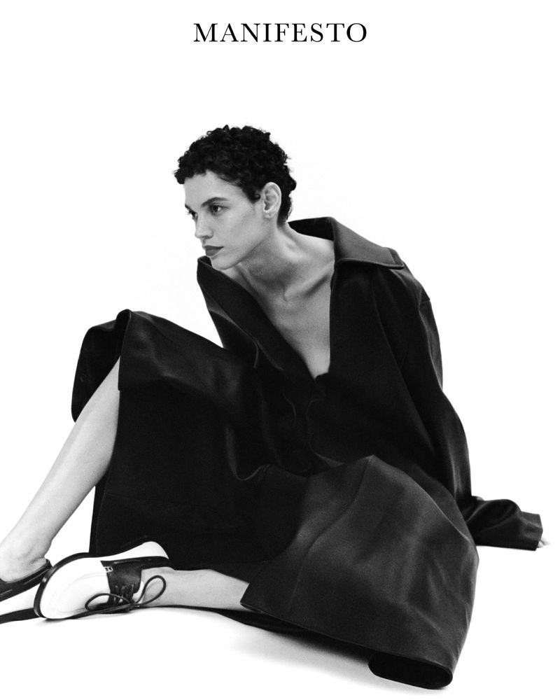 Model Kerolyn Soares sitting in a dark coat and two-tone shoes against a plain white background for Manifesto Magazine