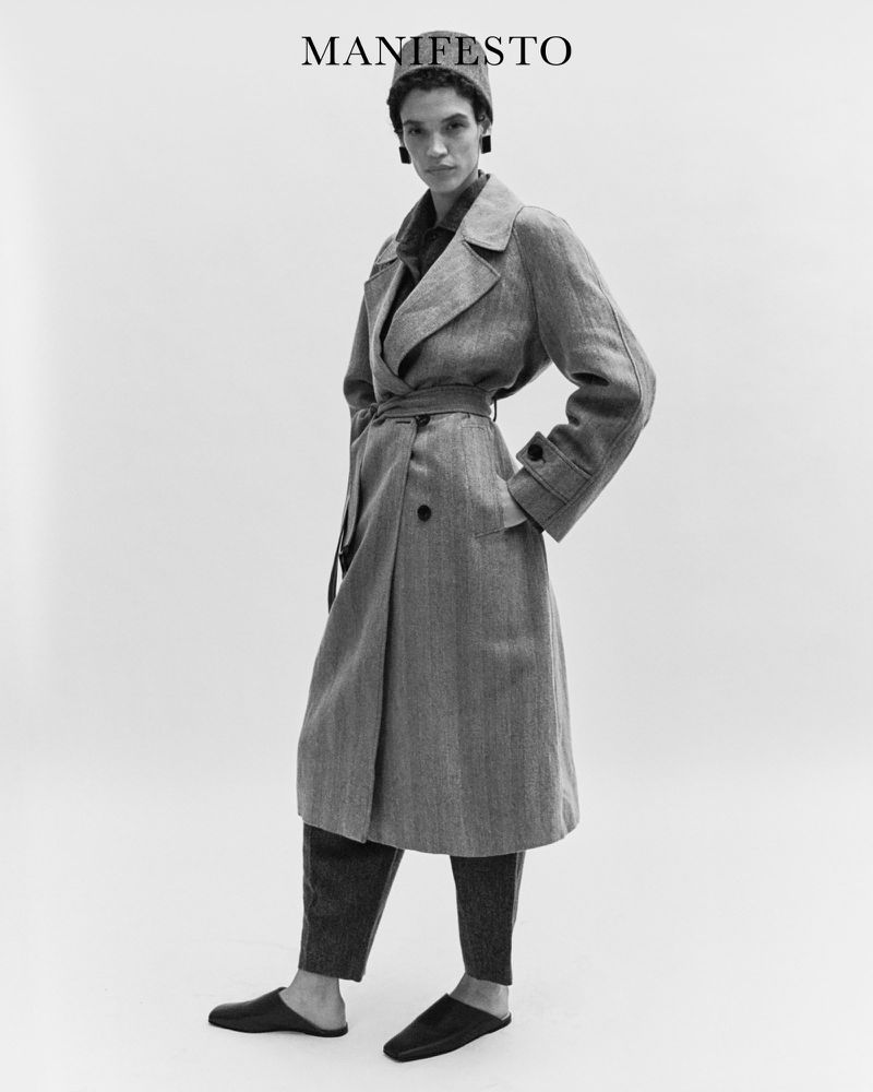Monochrome fashion shot of a woman in a long herringbone coat, trousers, and flat shoes, posing in a bright studio against a white backdrop.