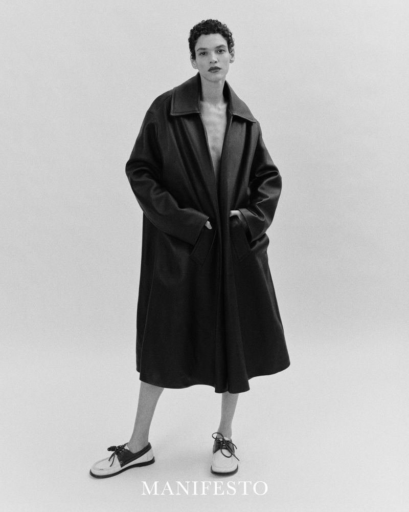 Full-length shot of a person with short hair wearing an oversized black coat and white shoes against a bright white background.