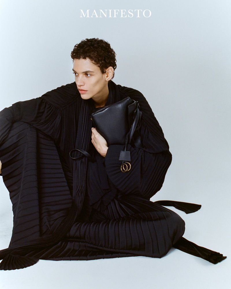 Fashion editorial shot of a slender male model in a draped black dress holding a black leather handbag against a seamless white backdrop.