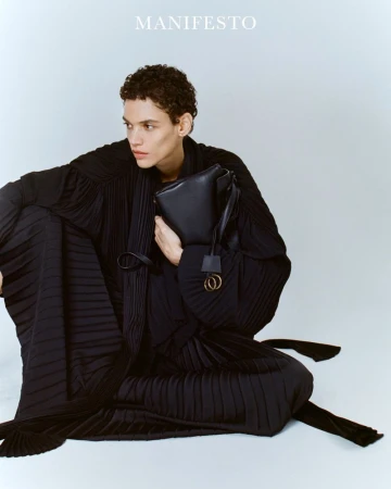 Fashion editorial shot of a slender male model in a draped black dress holding a black leather handbag against a seamless white backdrop.