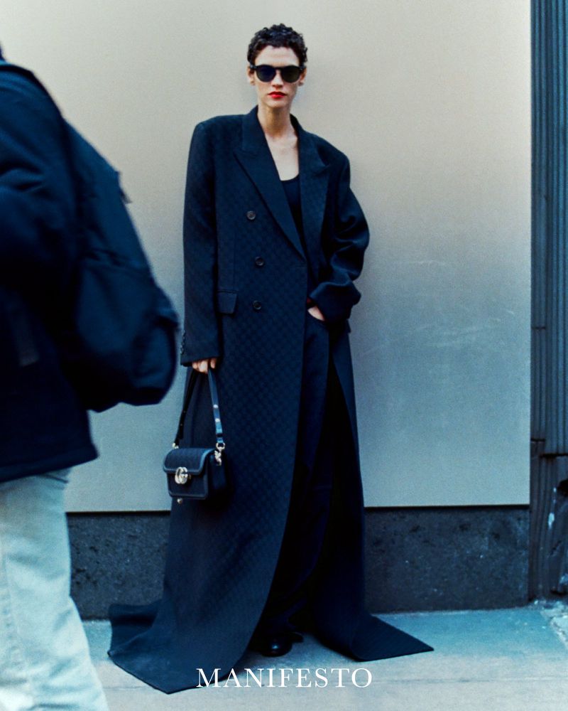 Fashion shot of a woman in a black overcoat and sunglasses, holding a purse, standing against a neutral wall in daylight.