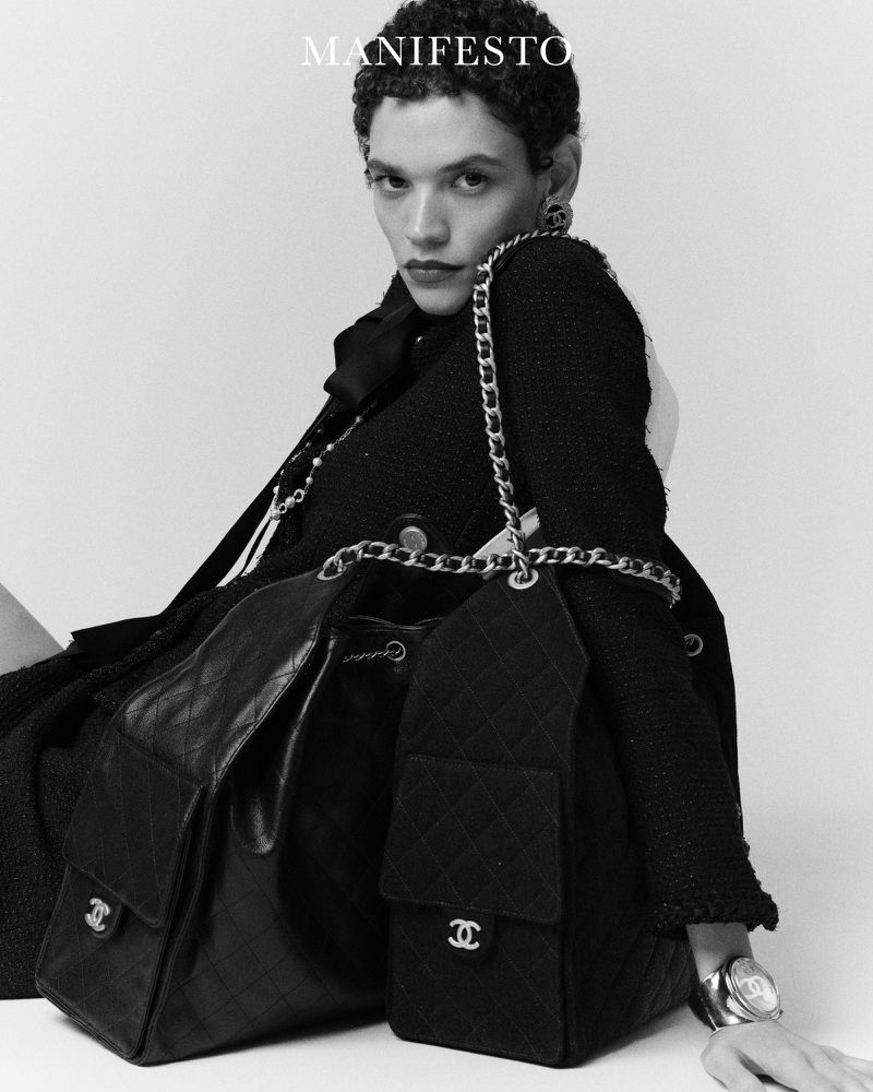 Fashion shot in black and white of a model wearing a Chanel jacket with chain straps on two Chanel bags, posing against a white background.