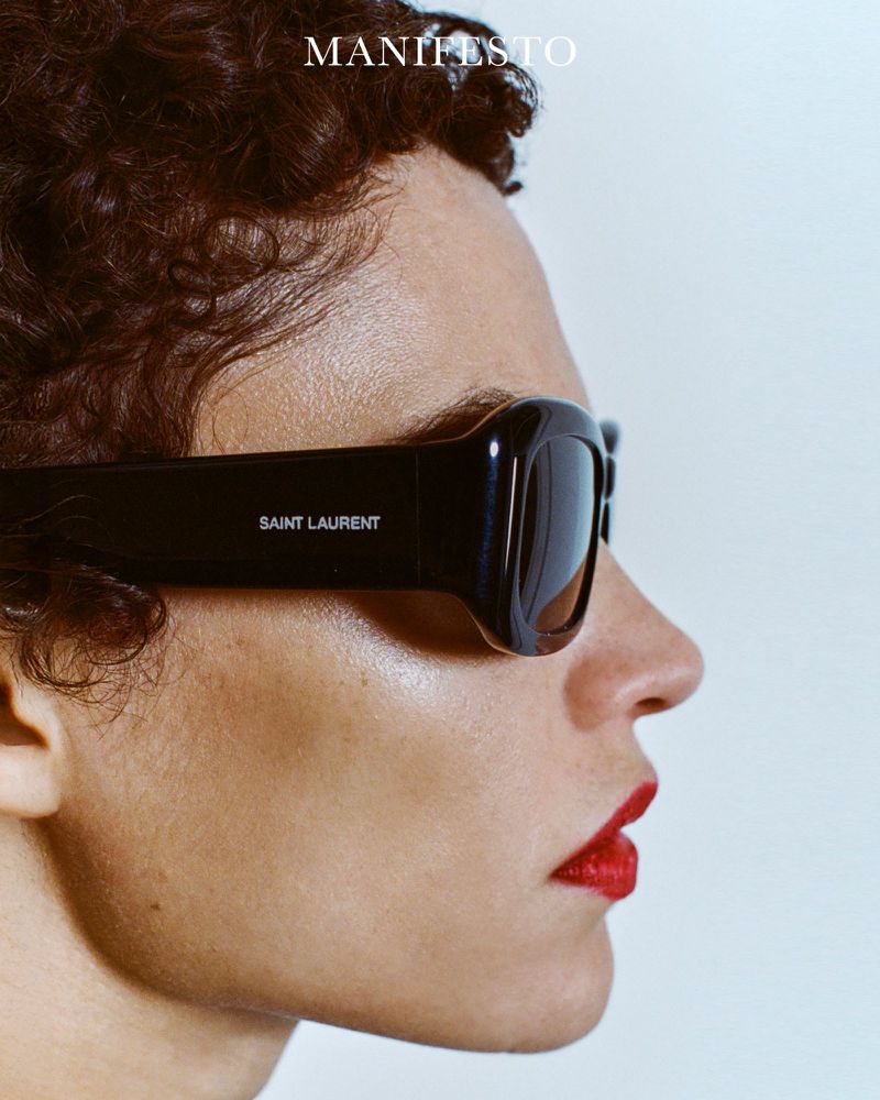 Close-up profile of a model wearing black Saint Laurent sunglasses and red lipstick against a plain white background for Manifesto Magazine.