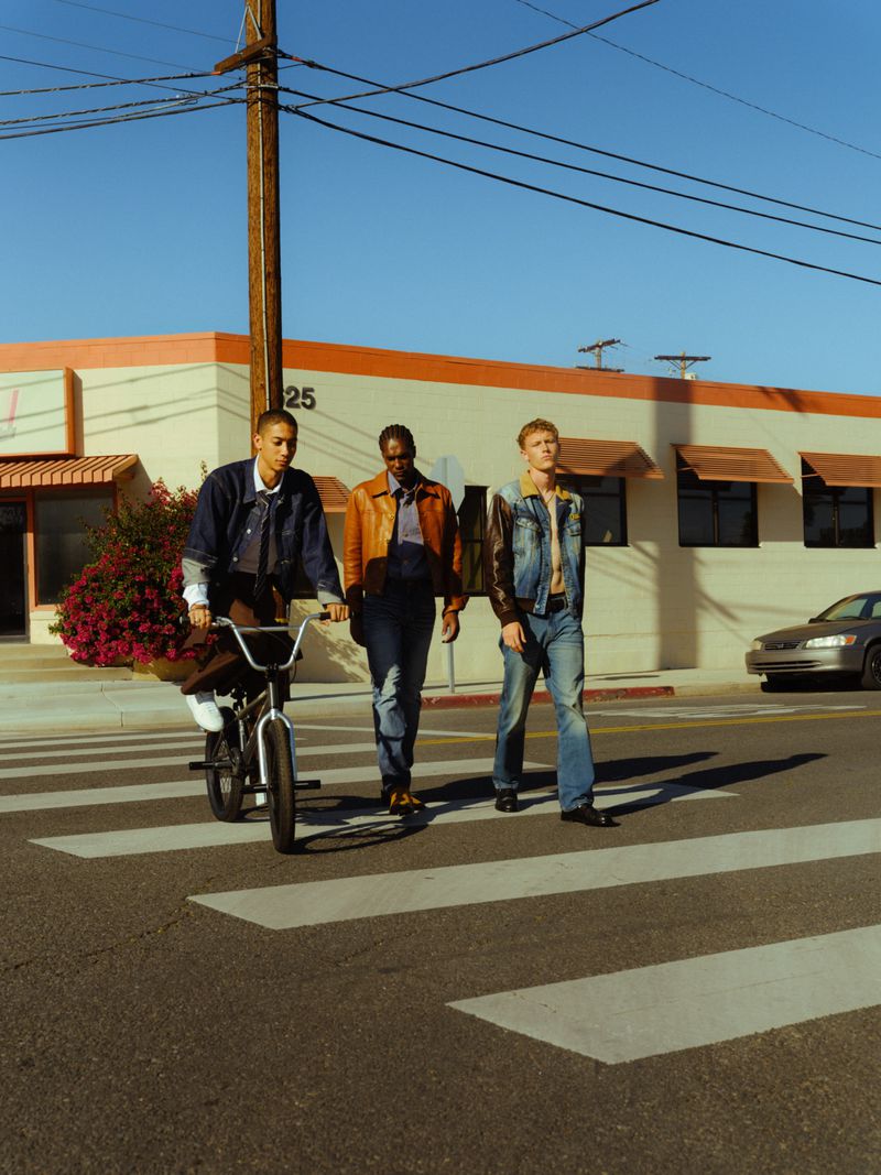 Three stylish men cross a street, one on a BMX bike, in a sunlit urban setting with a clear blue sky and a building in the background.