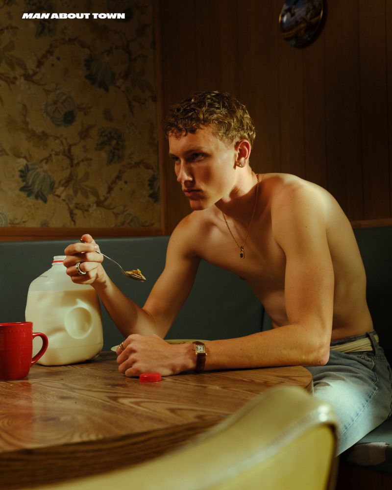 Shirtless man seated at a table with a milk jug, red mug, and fork in an indoor setting, with floral wallpaper in the background.