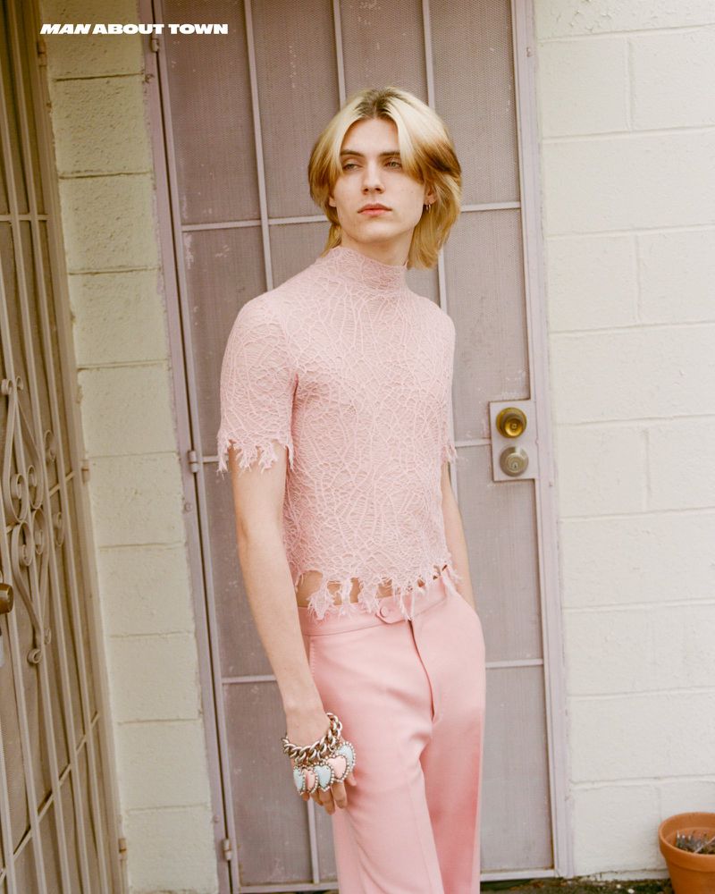 Young adult male poses in a pink lace shirt and matching pants, set against a weathered door and wall outdoors in soft, diffused light.