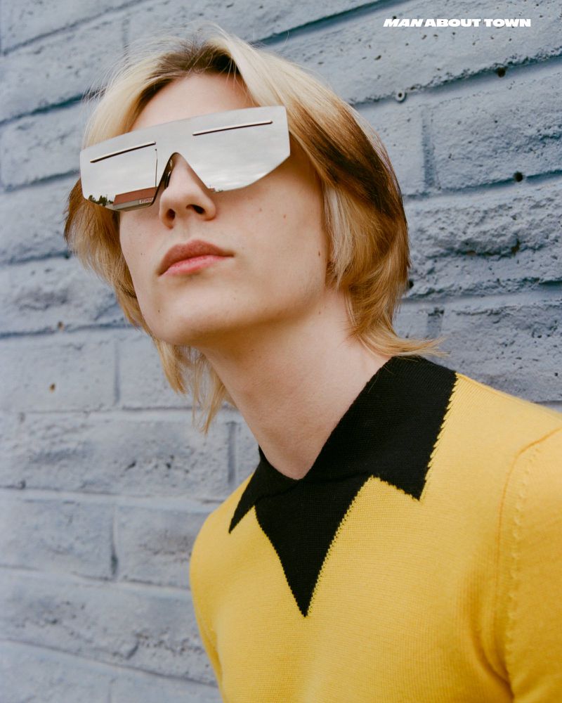 Close-up of a blond adult male wearing futuristic silver sunglasses and a yellow and black polo shirt against a brick wall.