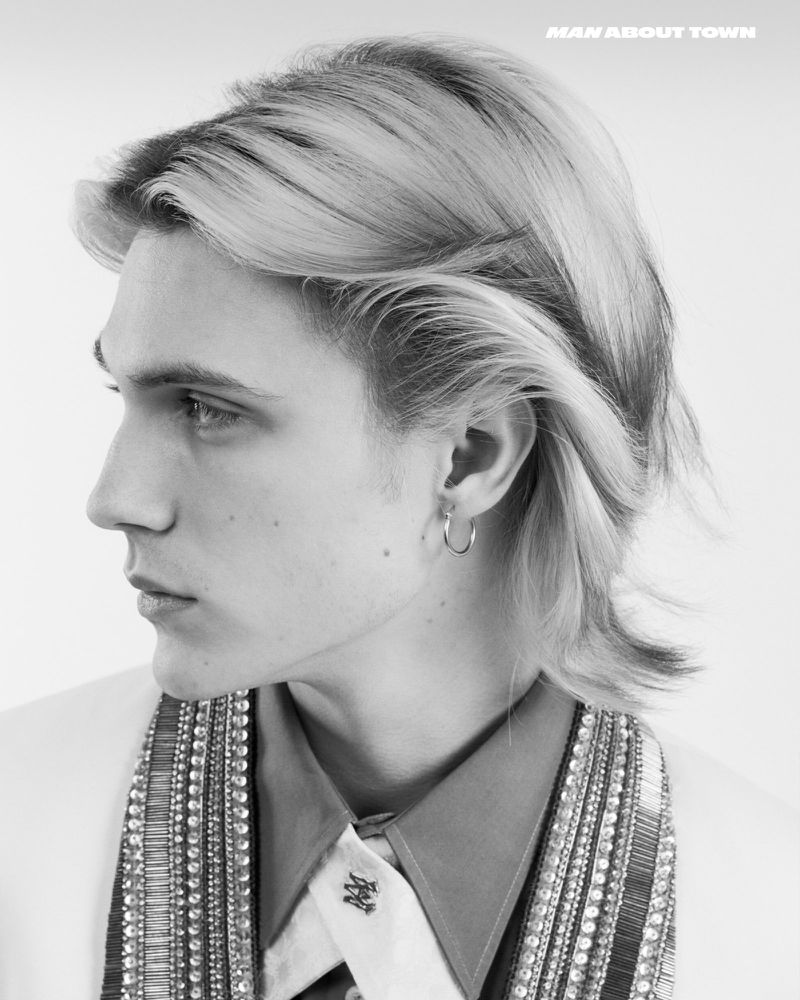 Black and white portrait of a young adult male with light blonde hair, wearing a collared shirt and beaded jacket, with a light backdrop.