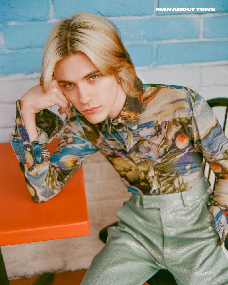 Portrait of a young adult with blonde hair wearing patterned button-down shirt and shiny pants, posing against blue brick wall.