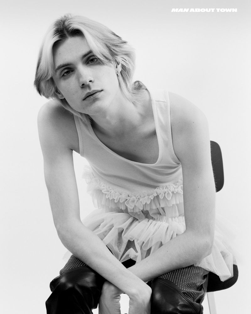 Portrait of a young adult with blond hair sitting on a chair against a white background, wearing a light-colored tank top and dark pants.