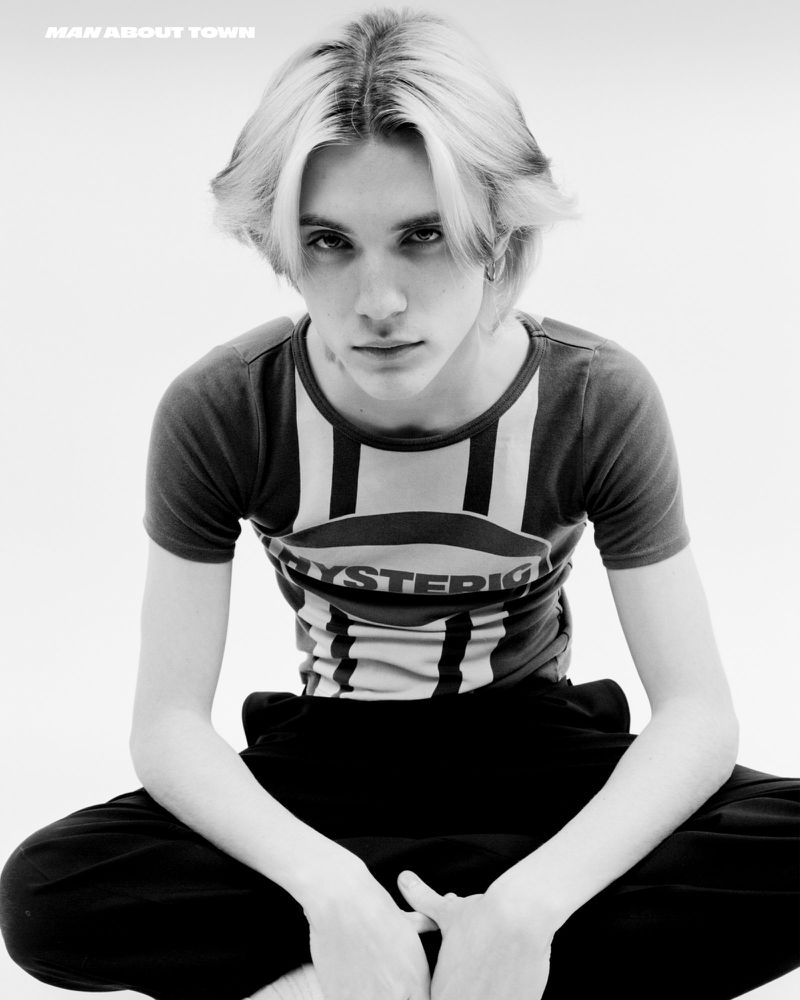 Monochrome portrait of a young adult with light blonde hair and a t-shirt with a graphic logo, seated against a bright white background.