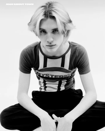 Monochrome portrait of a young adult with light blonde hair and a t-shirt with a graphic logo, seated against a bright white background.