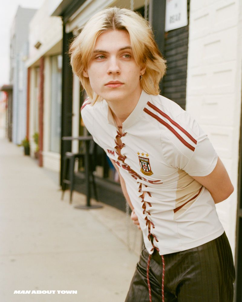 Young adult with blond highlighted hair in a soccer-style lace-up shirt in an urban outdoor setting near storefronts on a sunny day.