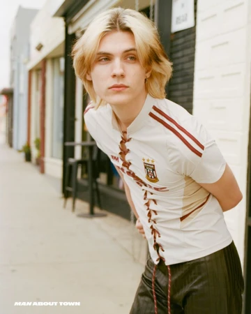 Young adult with blond highlighted hair in a soccer-style lace-up shirt in an urban outdoor setting near storefronts on a sunny day.