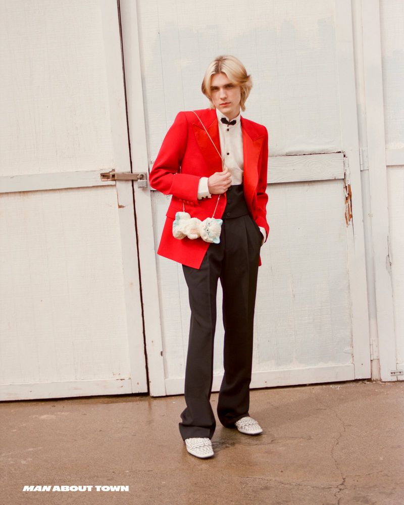 A fair-skinned male model with blond hair poses in a red suit jacket and black pants, holding a floral purse against a white wall.