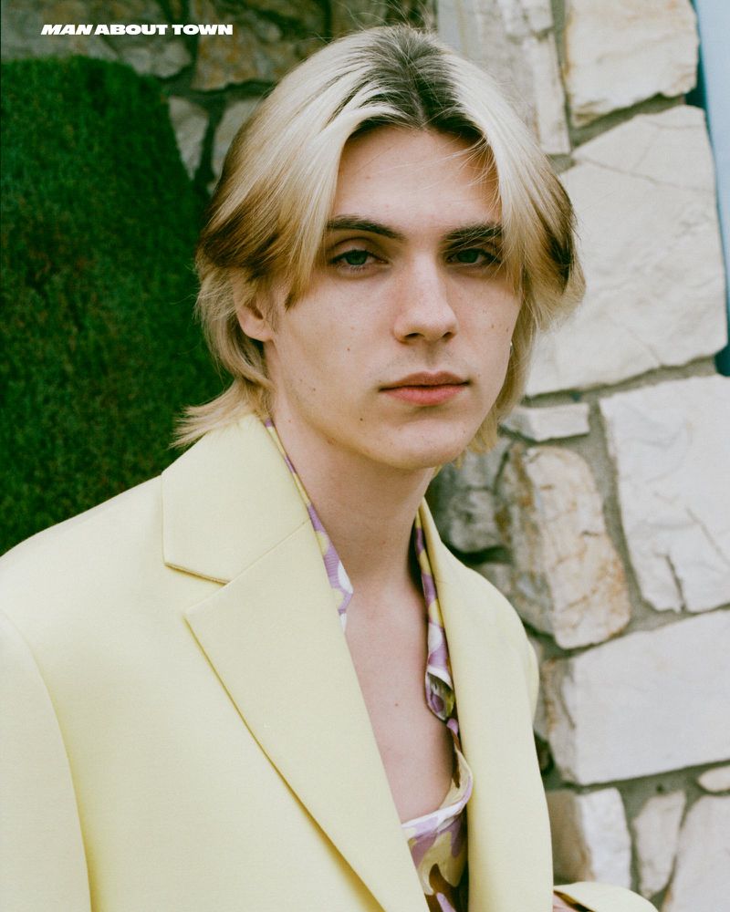 Portrait of a young man with blond and brown hair wearing a pale yellow suit jacket, with a light purple patterned shirt visible underneath.
