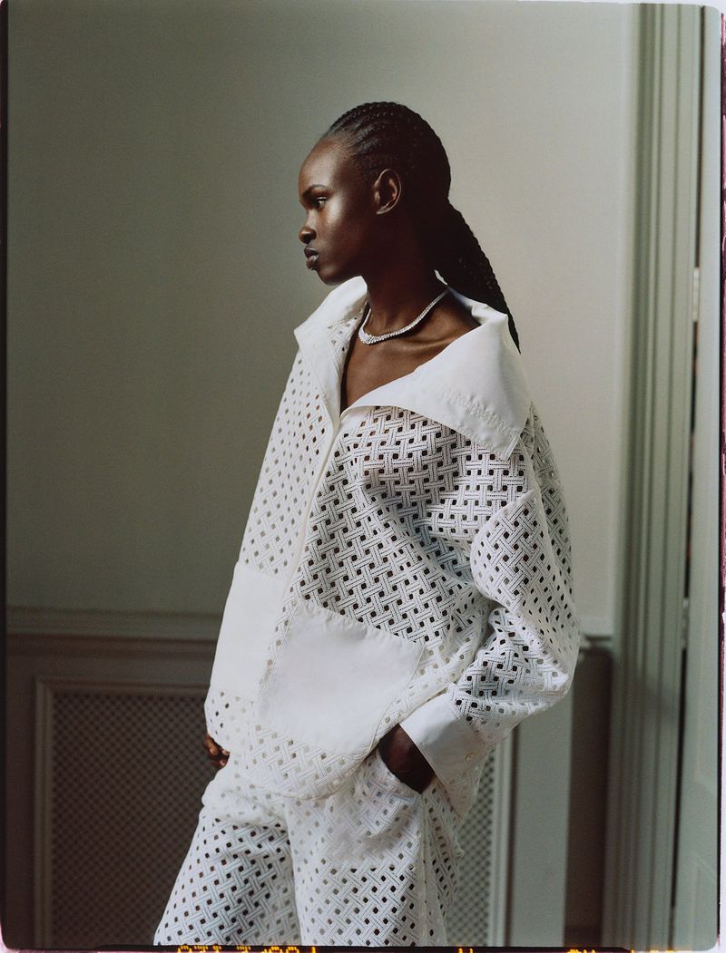 Portrait of a woman in an airy, white patterned jacket and matching pants, set against a simple light-toned indoor background.